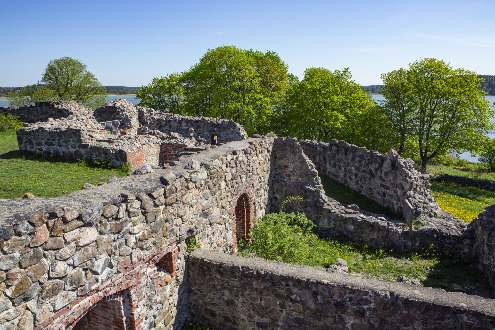Linnanraunioiden matalia kiviseiniä ja säilyneitä oviaukkoja. Raunioiden ympärillä vihreätä nurmikkoa, lehtipuita ja pensaita. Taustalla näkyy vettä ja sinistä taivasta. Kuva:Jari Kostet Linnanraunioiden matalia kiviseiniä ja säilyneitä oviaukkoja. Raunioiden ympärillä vihreätä nurmikkoa, lehtipuita ja pensaita. Taustalla näkyy vettä ja sinistä taivasta. Kuva:Jari Kostet