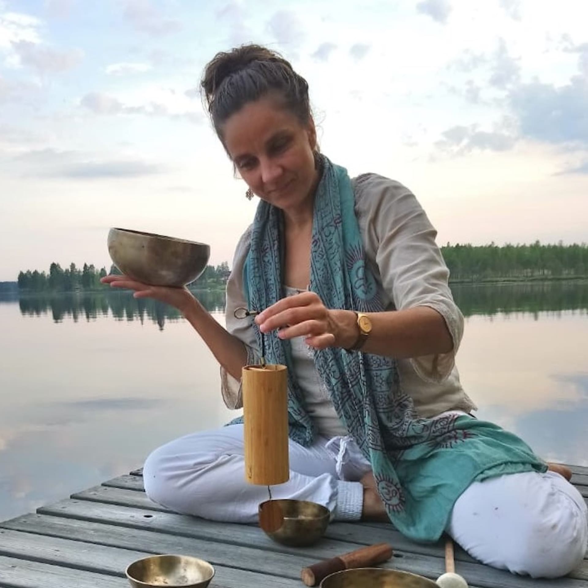 Woman framed by lake landscape playing sound bowls and koshi bells.