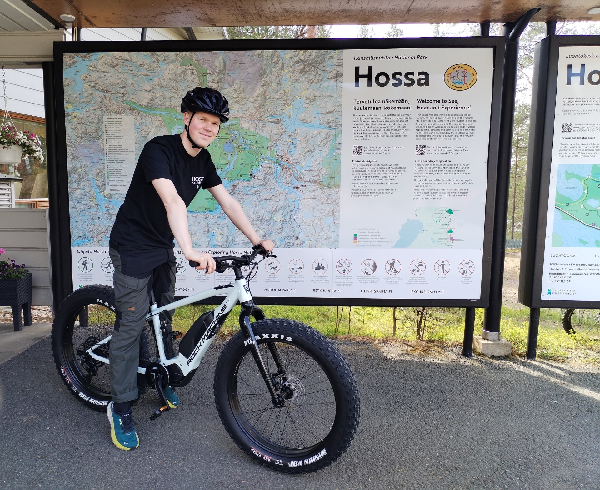 Pyöräilijä seisoo e-fatbiken kanssa Hossan kansallipuiston kartan edessä Hossan luontokeskuksen pihalla.