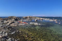 Kivikkoinen ranta, taustalla näkyy avomeri. Vesi on kirkasta, merenpohjassa näkyy kiviä. Punainen kajakki on vedetty rantaan. On kesä. Kuva: Timo Nieminen.