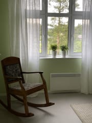 Huoneessa keinutuoli ikkunan vieressä. Ikkunalaudalla viherkasveja. Ikkunasta näkyy puita.