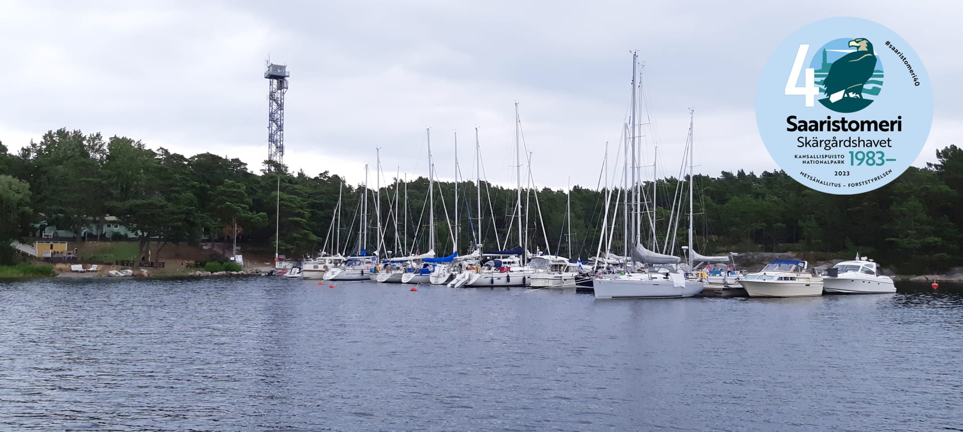 Pitkällä laiturilla on paljon veneitä. Rannalla  rakennus sekä korkea torni. The picture was taken from the water. There are many boats on the long pier. A building and a tall tower stand out on the beach. There are many trees on the beach.