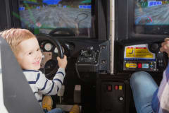 Pieni raitapaitainen poika istuu rallipelin ratin takana ja hymyilee olan takaa seuralaiselle jota ei näy kuvassa.