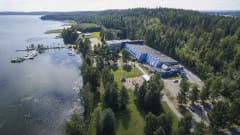 Iso sininen rakennus etualallaan pysäköintialue. Rakennus sijaitsee järven ja metsäisen harjun välissä. Rannalla puistoalue ja venlaiturit.