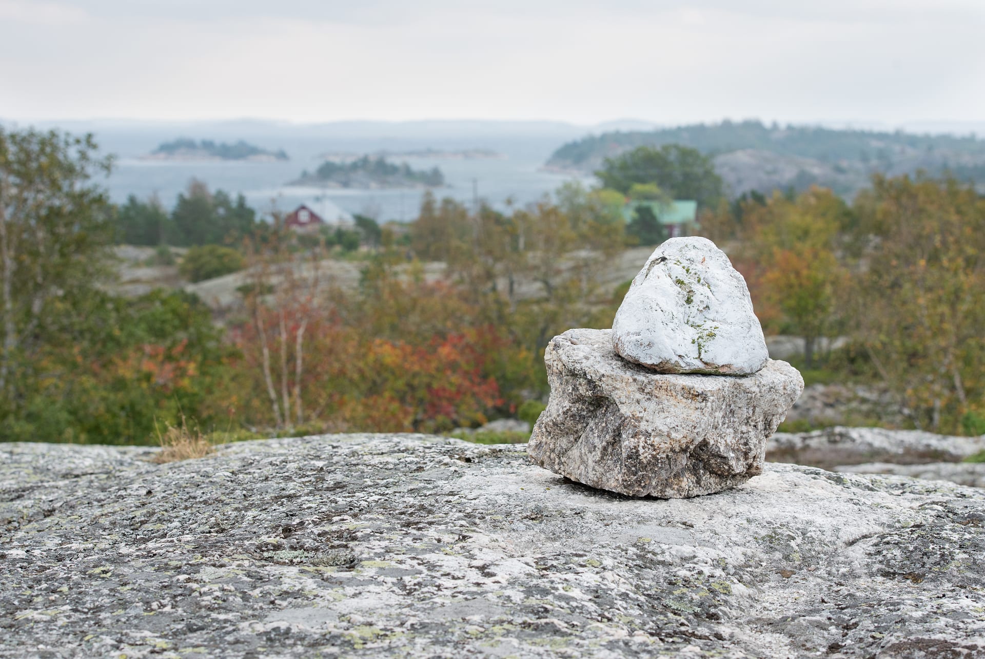 Kallion päällä kaksi valkoiseksi maalattua kiveä päällekkäin. Takana siintää kylään kuuluvia rakennuksia. In the right foreground, on top of a rock, two white painted stones on top of each other. 