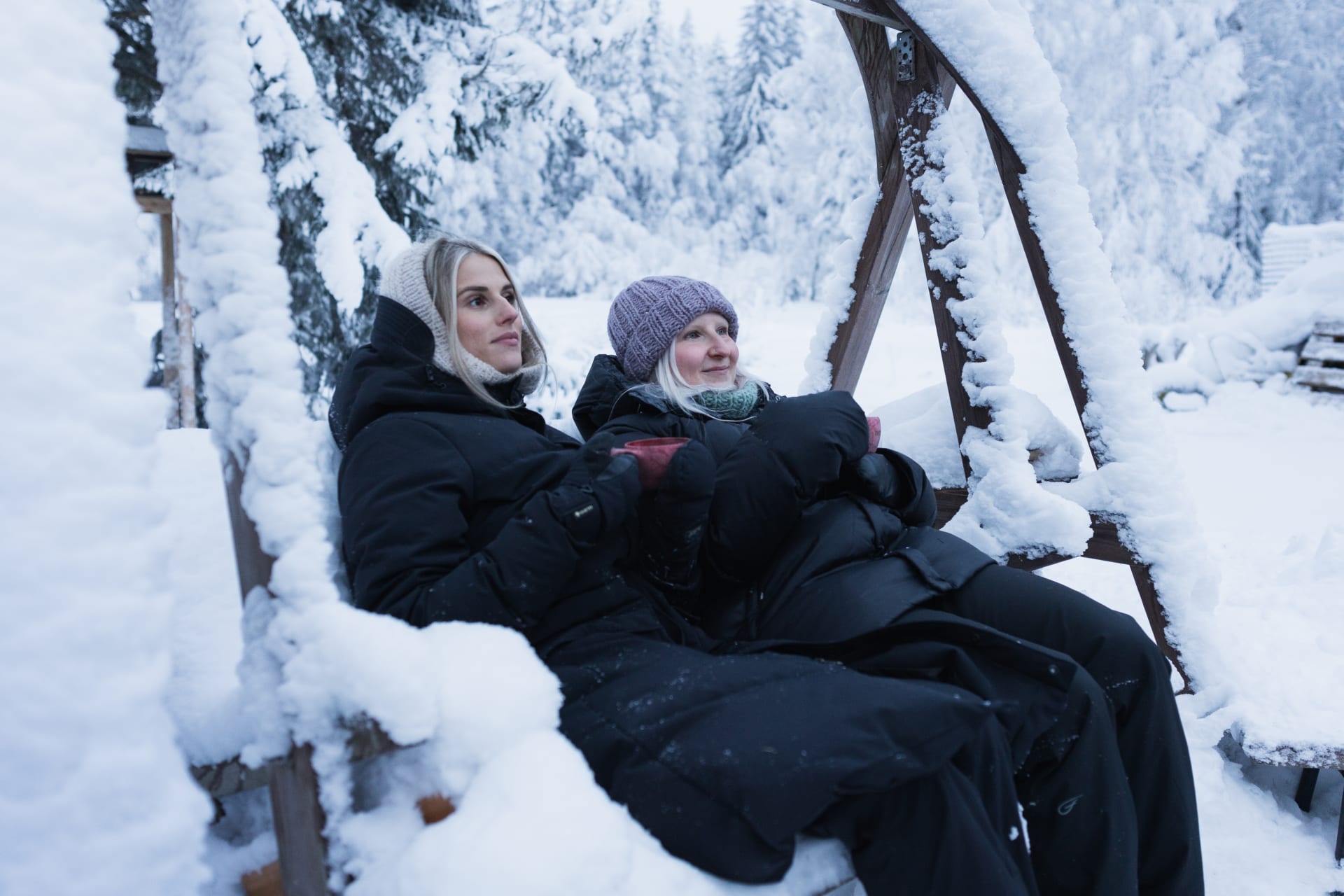 Kaksi muorta naista talviasuissaan istuu kiikussa, maisema on luminen.