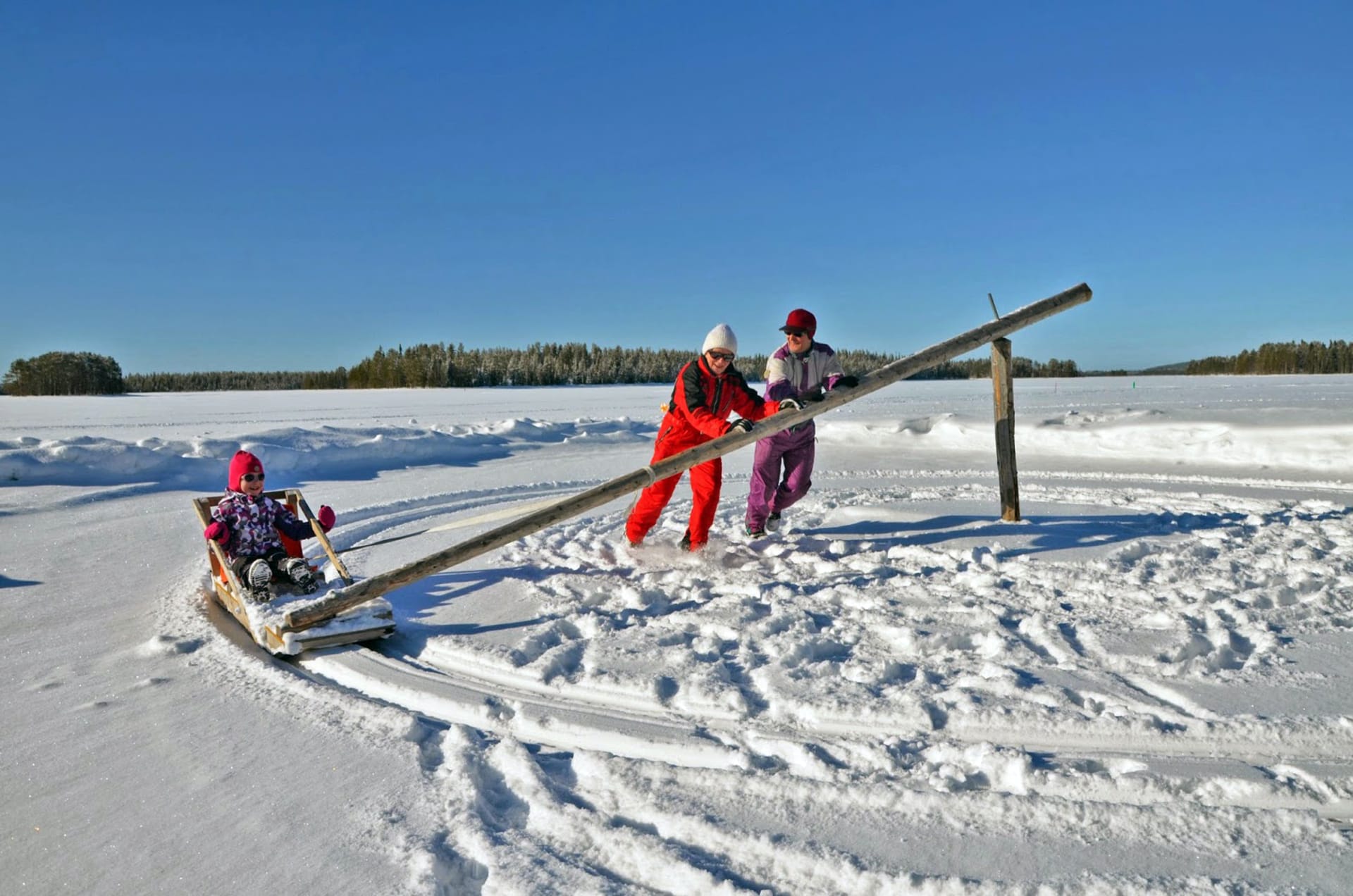 Hoijakka, the traditional merry-go-round sled, circles on sunny lake ice, with parents pushing to speed up for the rejoicing child in the sled.