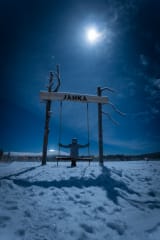 A person sits on a wooden swing beneath the Jänkä sign at night, illuminated by moonlight and stars in a snowy Lapland landscape.