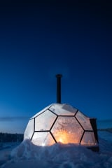 A glass fire shelter glows from within on a snowy hillside at Jänkä, set against a dark blue night sky and winter forest.