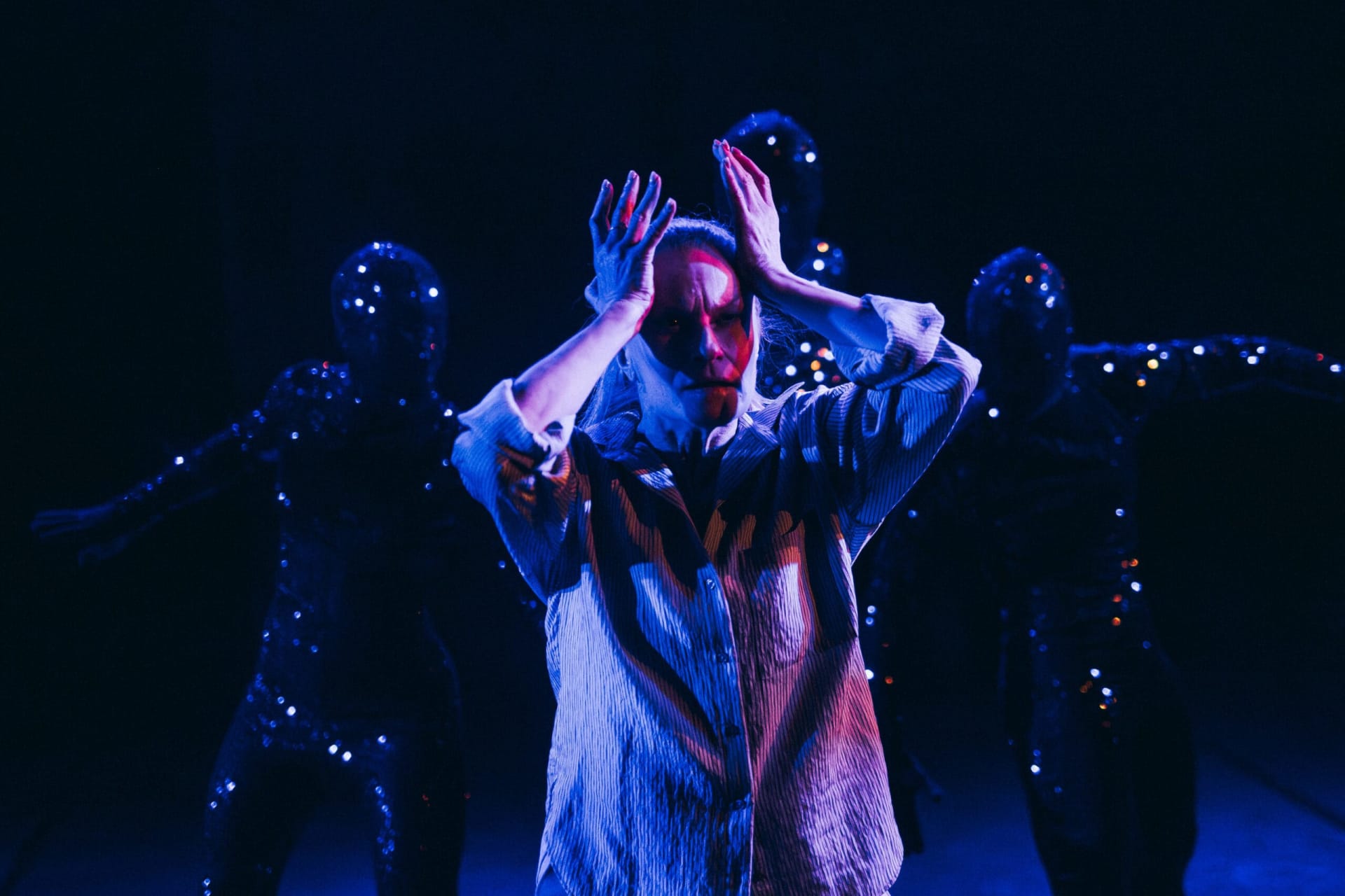 A dancer stands in blue and purple lighting with her hands around her face. Four dancers in dark costumes with small points of light stand in the background. The image is from Aurinkobaletti’s dance production Lumen.