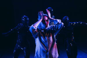 A dancer stands in blue and purple lighting with her hands around her face. Four dancers in dark costumes with small points of light stand in the background. The image is from Aurinkobaletti’s dance production Lumen.