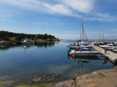 Oikealla laituri, jossa on kiinni purje- ja moottoriveneitä. Edessä kaksi ruskettunutta veneilijää. Vasemmalla toinen laituri, jossa on veneitä kiinni. Laiturin takana on kallioita ja metsää. Meri on tyyni, taivaalla poutapilviä. Kuva: Kirsi Nikkola.