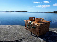 Punottu, nelikulmainen eväskori rantakalliolla auringonpaisteessa, korin kannella kaksi puista juomakuppia, kuksaa. Taustalla tyyni sininen meri ja pilvetön taivas. Kuva: Matti Piirainen.