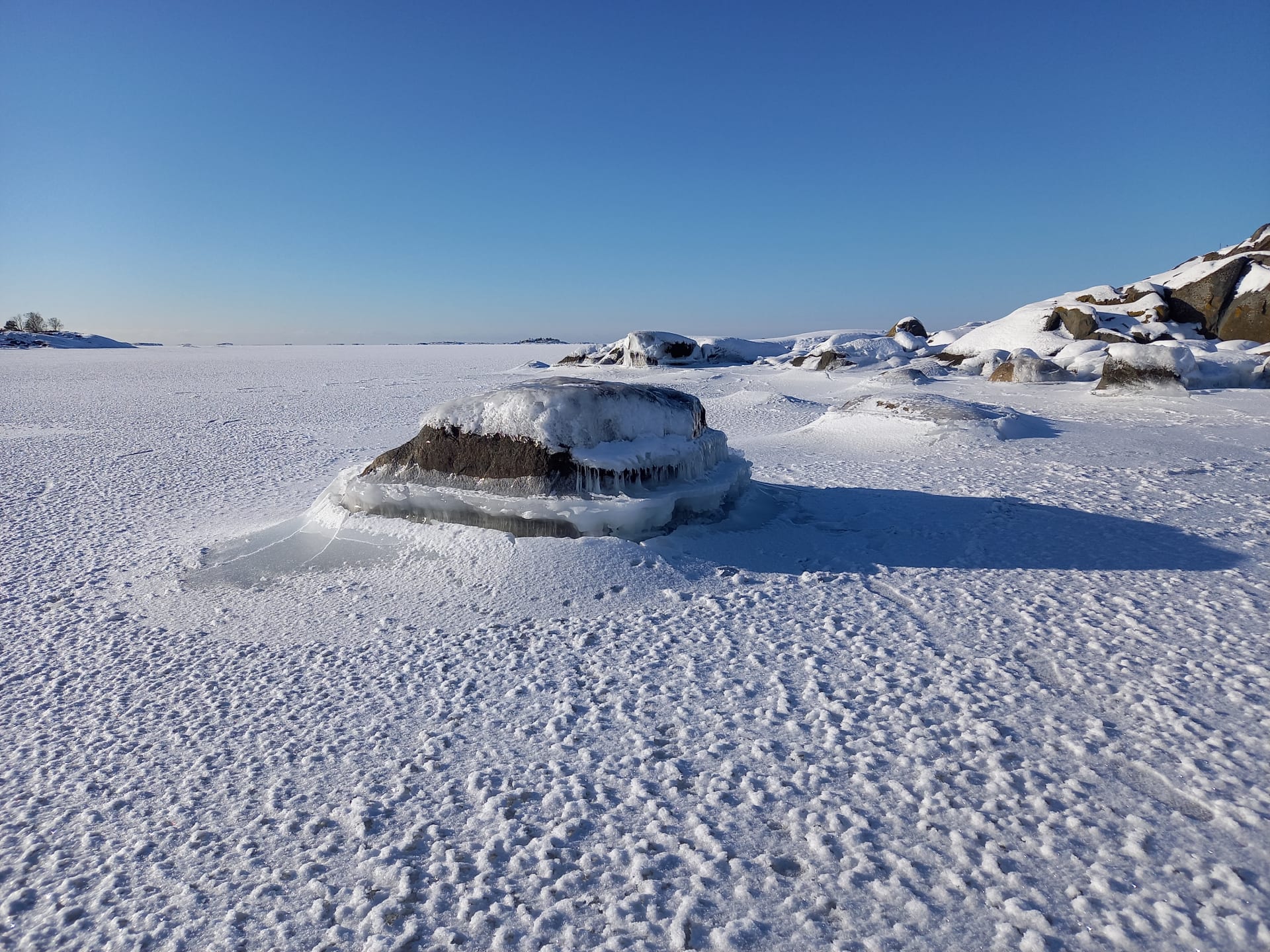 Lumen peittämä ja jään ympäröimä rantakivi ja jäätynyttä merenpintaa aurinkoisena talvipäivänä. Kuva: Matti Piirainen.