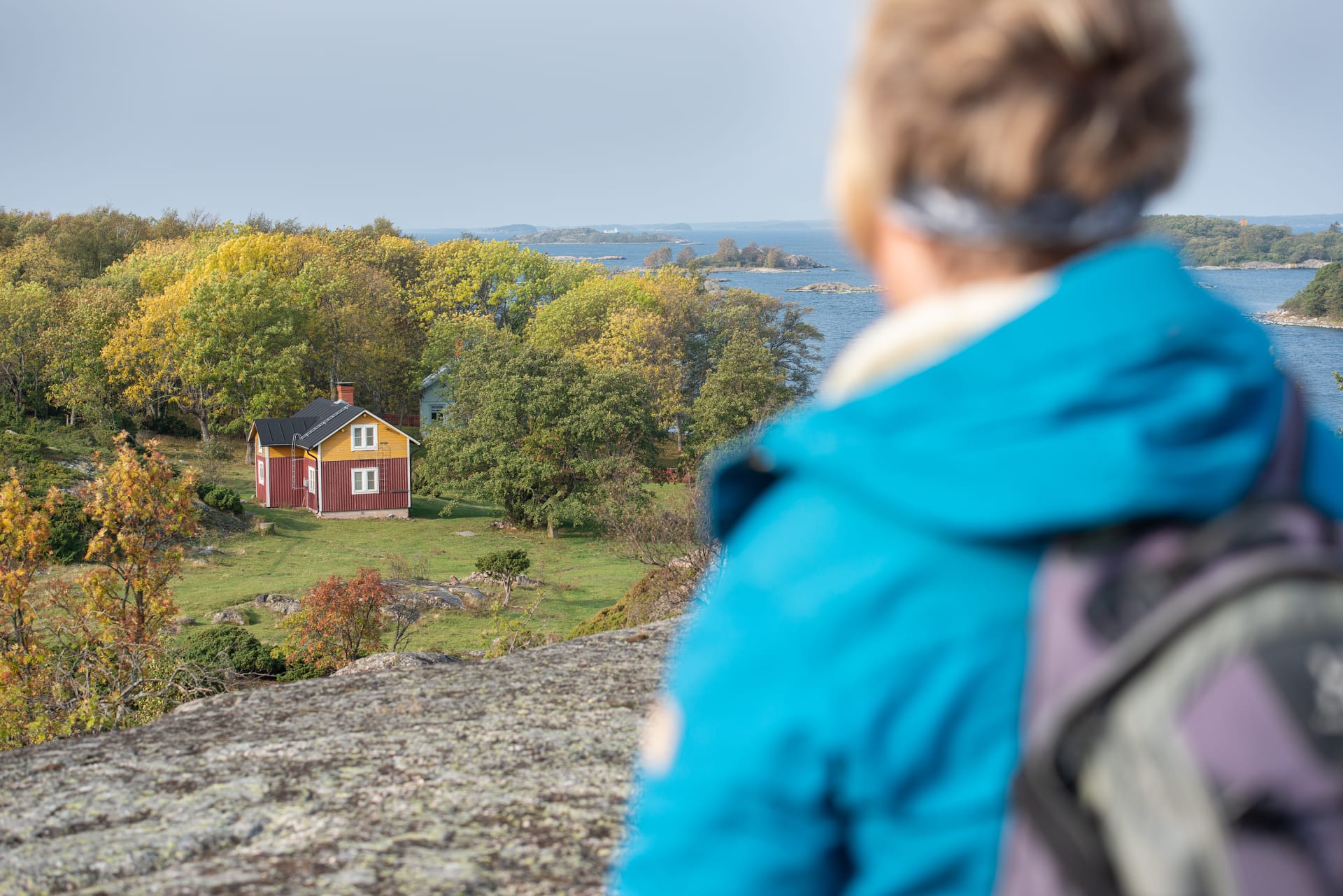 Nainen seisoo puisella riippusillalla ja katsoo maisemaa. Ympärillä kalliota ja puustoa. Kauempana siintää meri. A woman stands on a wooden suspension bridge and looks at the landscape. Rocks and trees all around. In the distance is the sea.