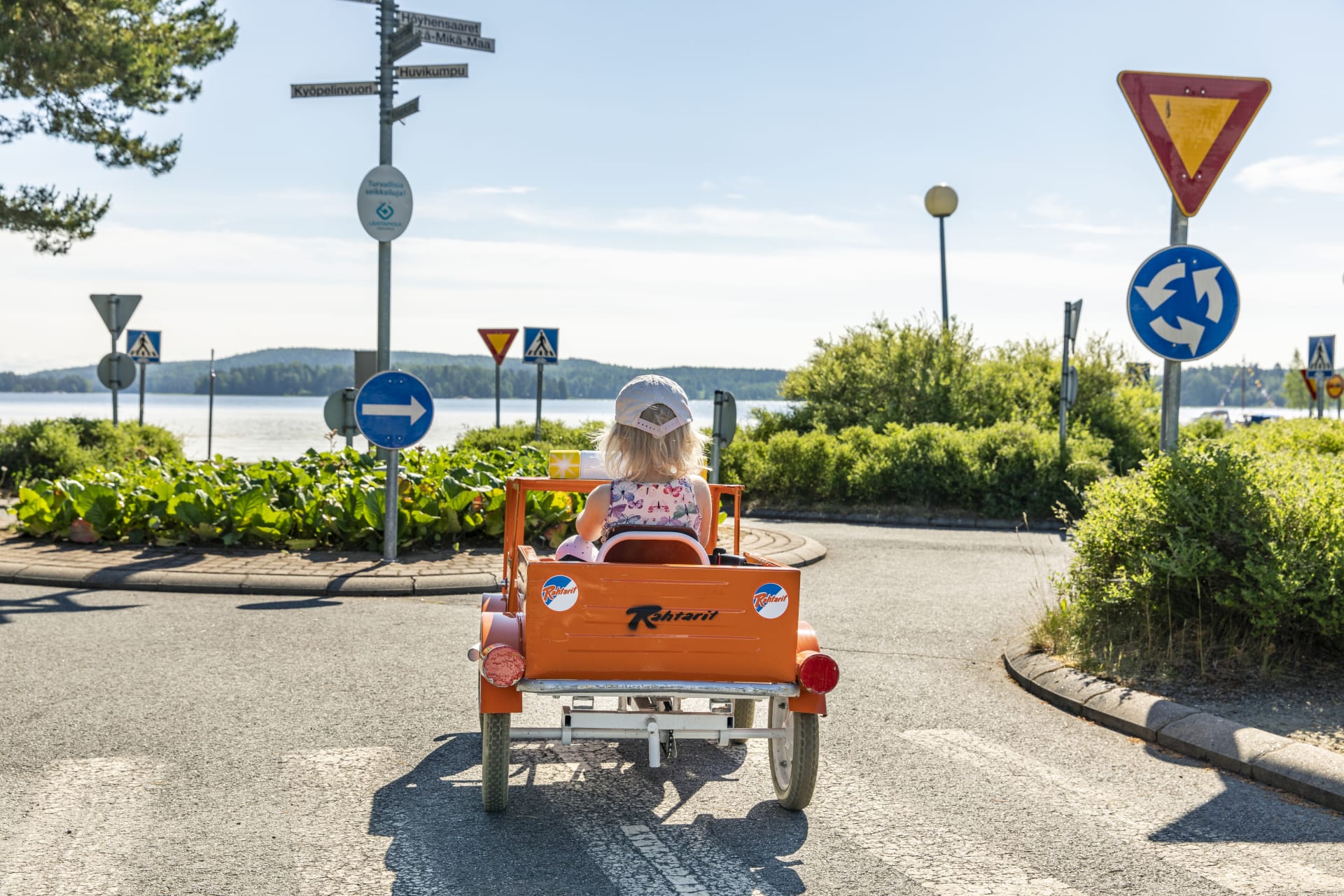Lippalakkipäinen tyttö oranssissa polkuautossa ajaa liikenneympyrään. Taustalla järvi.