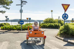 Lippalakkipäinen tyttö oranssissa polkuautossa ajaa liikenneympyrään. Taustalla järvi.