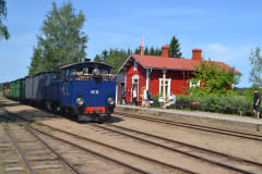 Blue steam locomotive in front of red wooden Minkiö station building under nice blue summer sky.