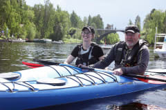A woman and a man are next to each other in single kayaks, smiling at the camera in the Oravi Canal on Lake Saimaa