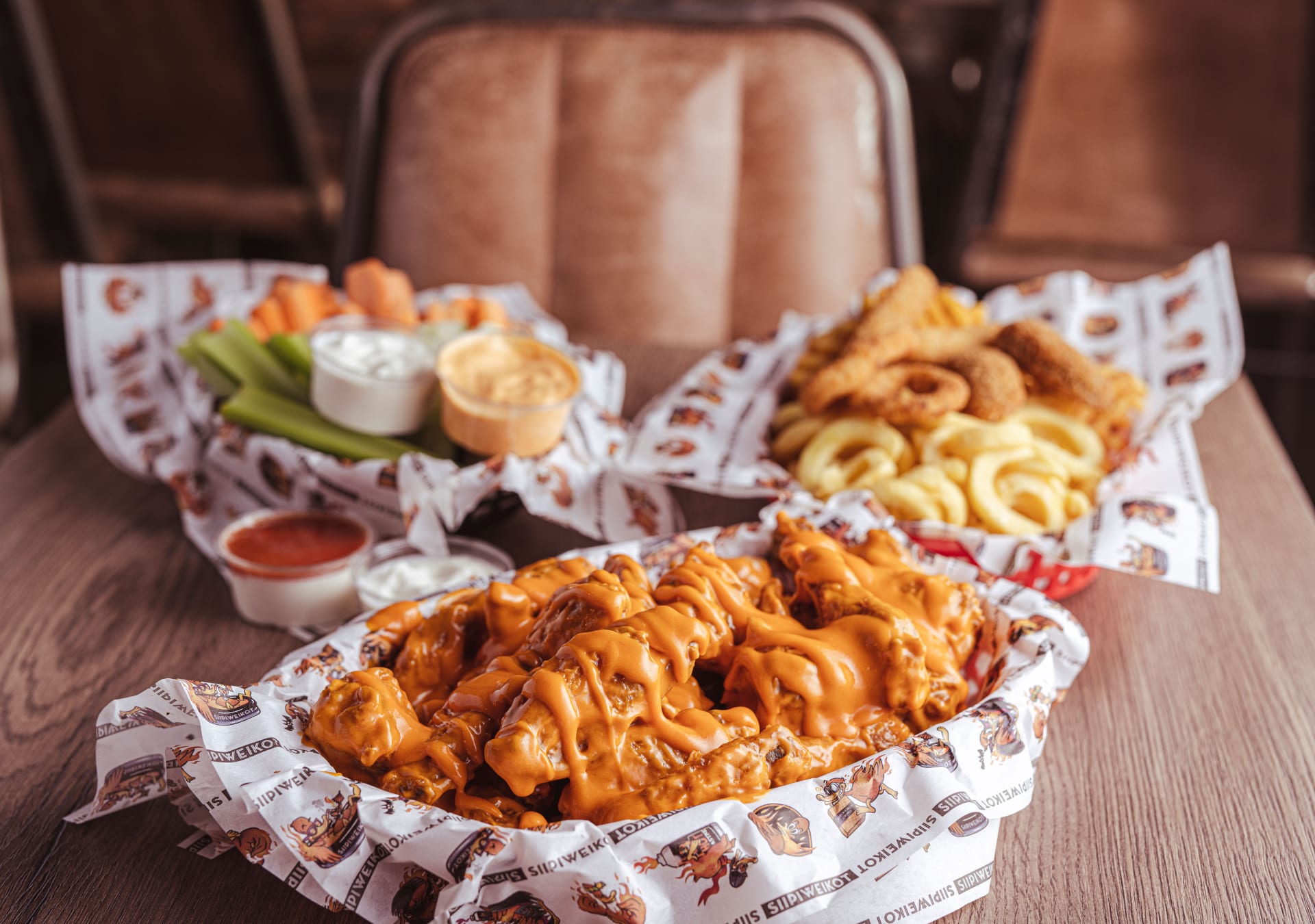 Crispy chicken wings coated in orange buffalo sauce on a tray. Celery and carrot sticks with ranch dip on the side. Onion rings and fries in a basket in the background. Served on a wooden table in a restaurant setting.