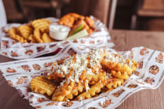 Crispy waffle fries topped with buffalo sauce and crumbled blue cheese. Chicken wings with dip and vegetable sticks in the background. Served on a wooden table in a restaurant setting.