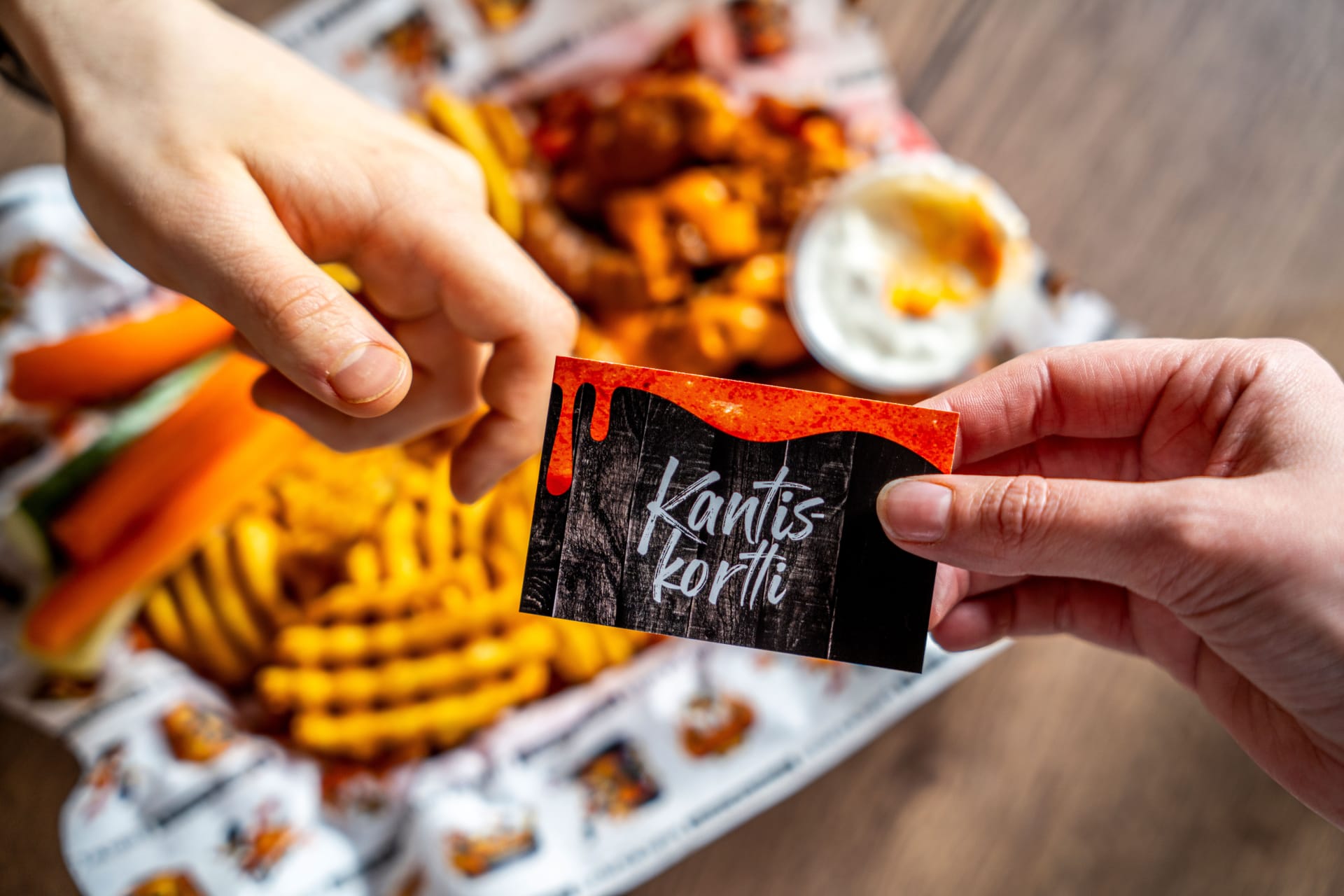 Hands holding a loyalty card with the text “Kantiskortti.” Chicken wings, waffle fries, and vegetable sticks with dip in the background.