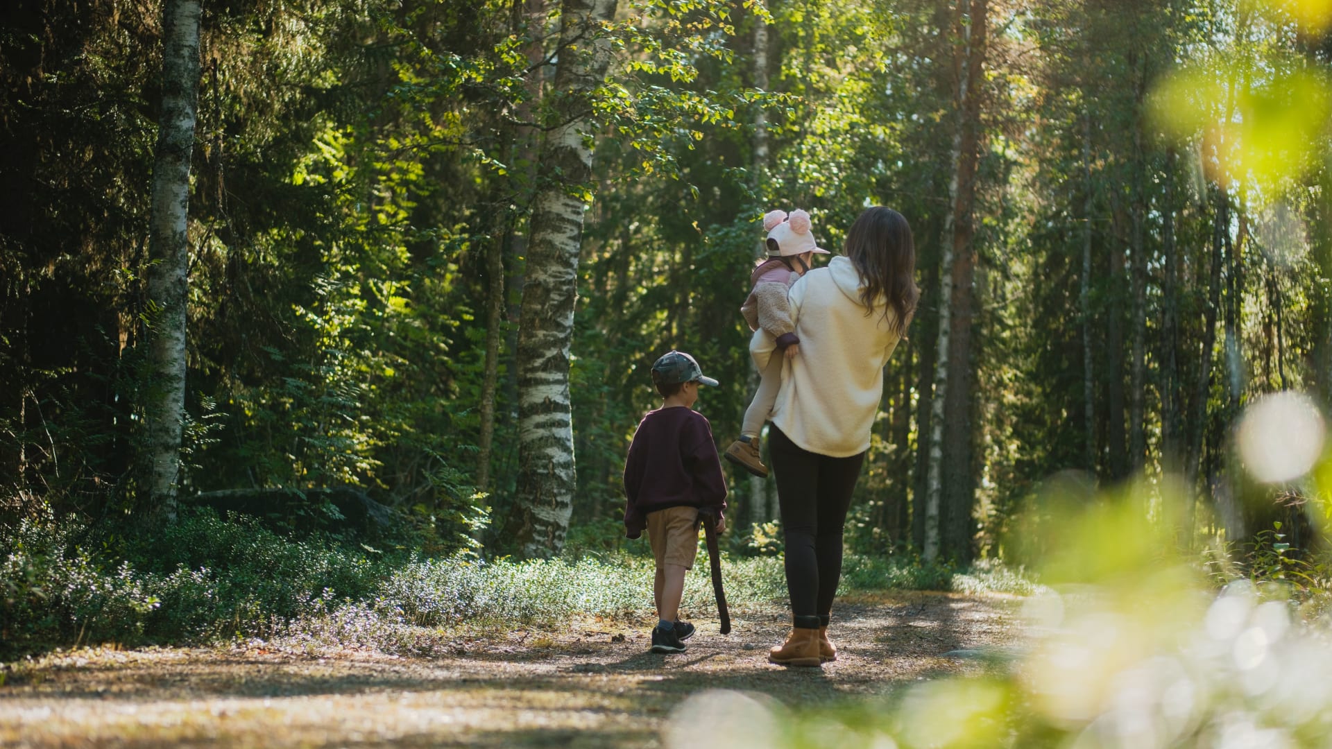 Mother carrying her daughter and son walking next to them in the luscious forest.
