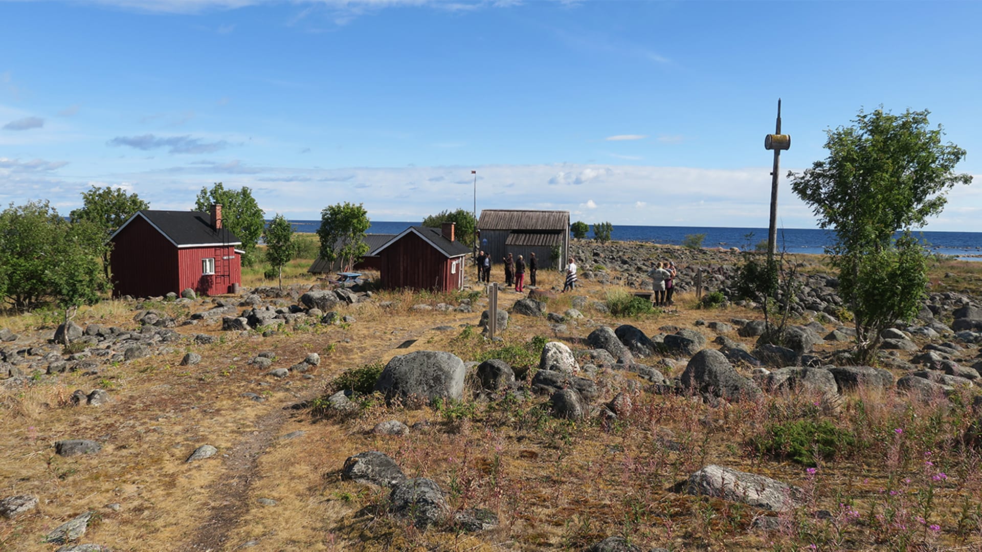 Kivikkoinen saari, jossa ryhmä ihmisiä. Pihassa vanhoja rakennuksia ja tynnyrisalko. Merenranta taustalla. / A rocky island with a group of people. In the yard old wooden buildings and a barrel stove. The sea shore in the back. Kuva: Irja Leppänen.