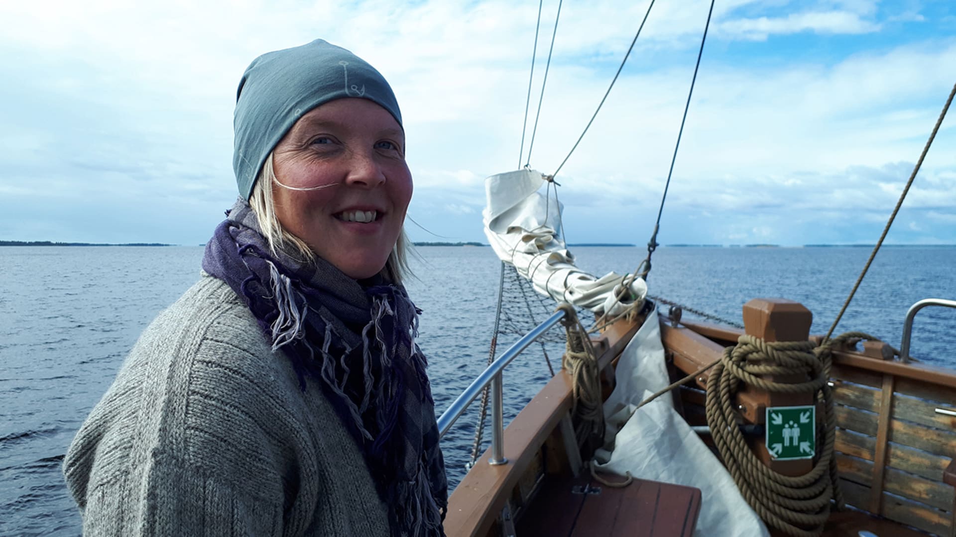 Hymyilevä henkilö laivassa. Taustalla näkyy laivan perä purjeineen ja köysineen. / Smiling person on board. The stern of the ship with sails and ropes can be seen in the background. Kuva: Heli Suurkuukka