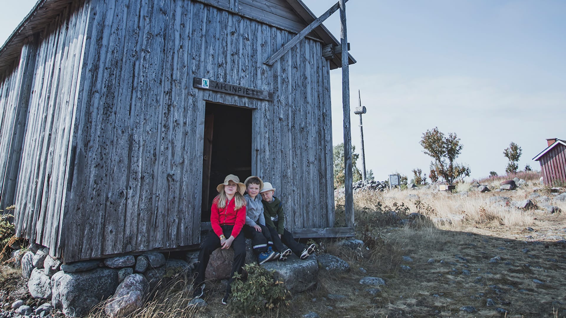 Kolme hymyilevää lasta istumassa kalamajan portaalla. /  Three smiling children sitting on the steps of a fishing house. Kuva:Maarit Vaahteranoksa