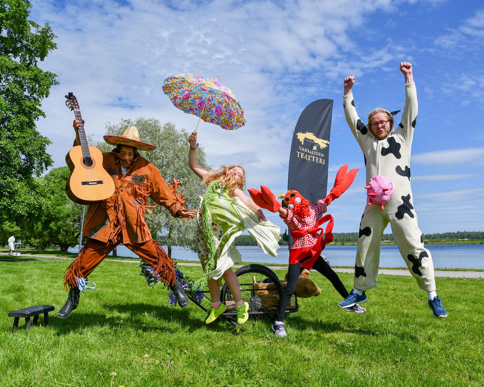 Joyful children's street theatre group consists of four actors. A mexican guitar player, a ladylike girl, a girl in crab costume and a man in cow costume are jumping with their hands up.