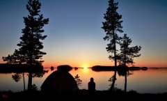 Retkeilijä ja teltta rannalla kesäisessä Etelä-Konneveden kansallispuistossa. Taustalla peilityyni järvimaisema ja auringonlasku. Kuva: Janita Juvakka