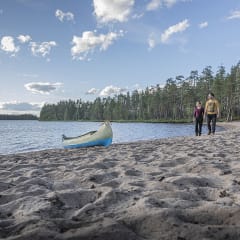 Kaksi ihmistä kävelee hiekkarannalla, aivan veden rajassa. Rannassa on kanootti. Kuva: Merja Paakkanen