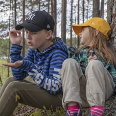 Kaksi lasta istuu maassa männyn juurella . Toisella on kädessään hyönteinen. Kuva: Merja Paakkanen
