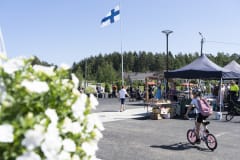 Vesilahden Kirkonkylän tori ja liehuva Suomen lippu sekä torilla kävijöitä - ©