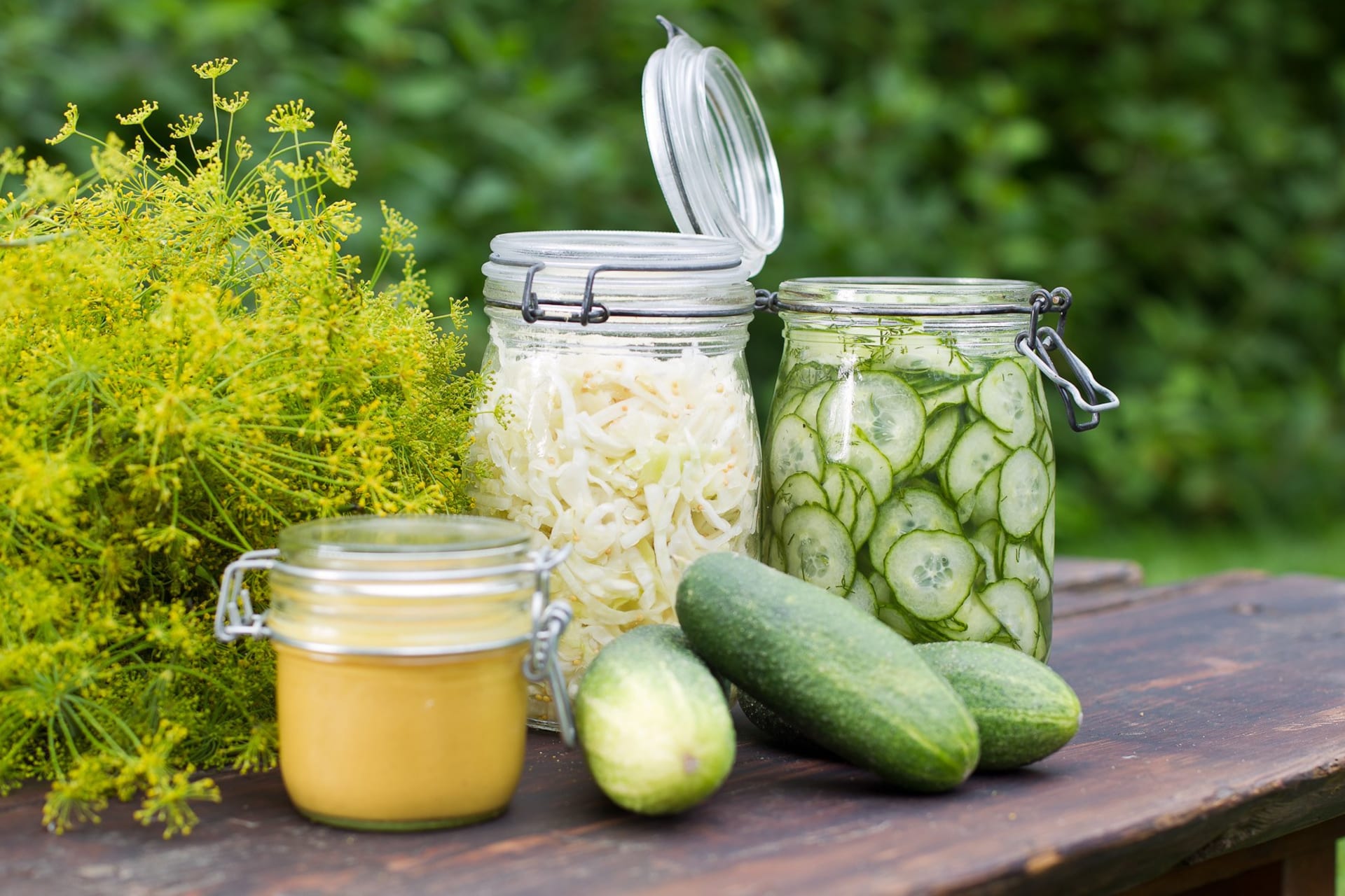 Three glass jars filled with preserved vegetables. Three cucumbers in the front. 