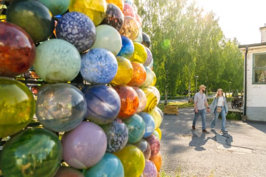 A couple walking in the Nuutajärvi Glass Village. In front of the picture is a colourful glass ball tower.