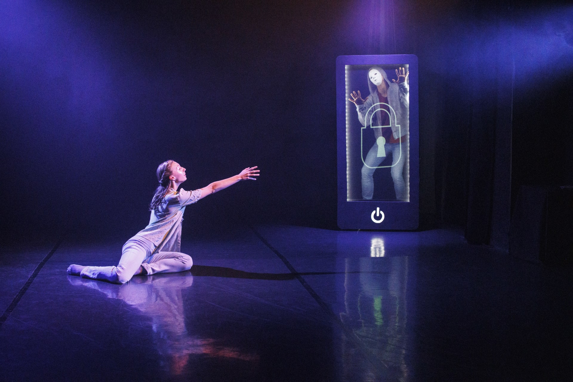 Tyttö istuu lattialla ojentaen kättään kohti puhelinta, jonka sisällä on toinen henkilö Tyttö istuu lattialla ojentaen kättään kohti puhelinta, jonka sisällä on toinen henkilö