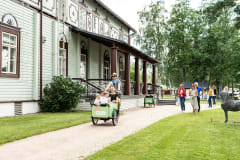 Mies kuljettaa lapsia polkupyörän etukärryssä Taidekeskus Salmelan edessä. A cycling man having children in the front carriage of a bicycle in front of Art Center Salmela.