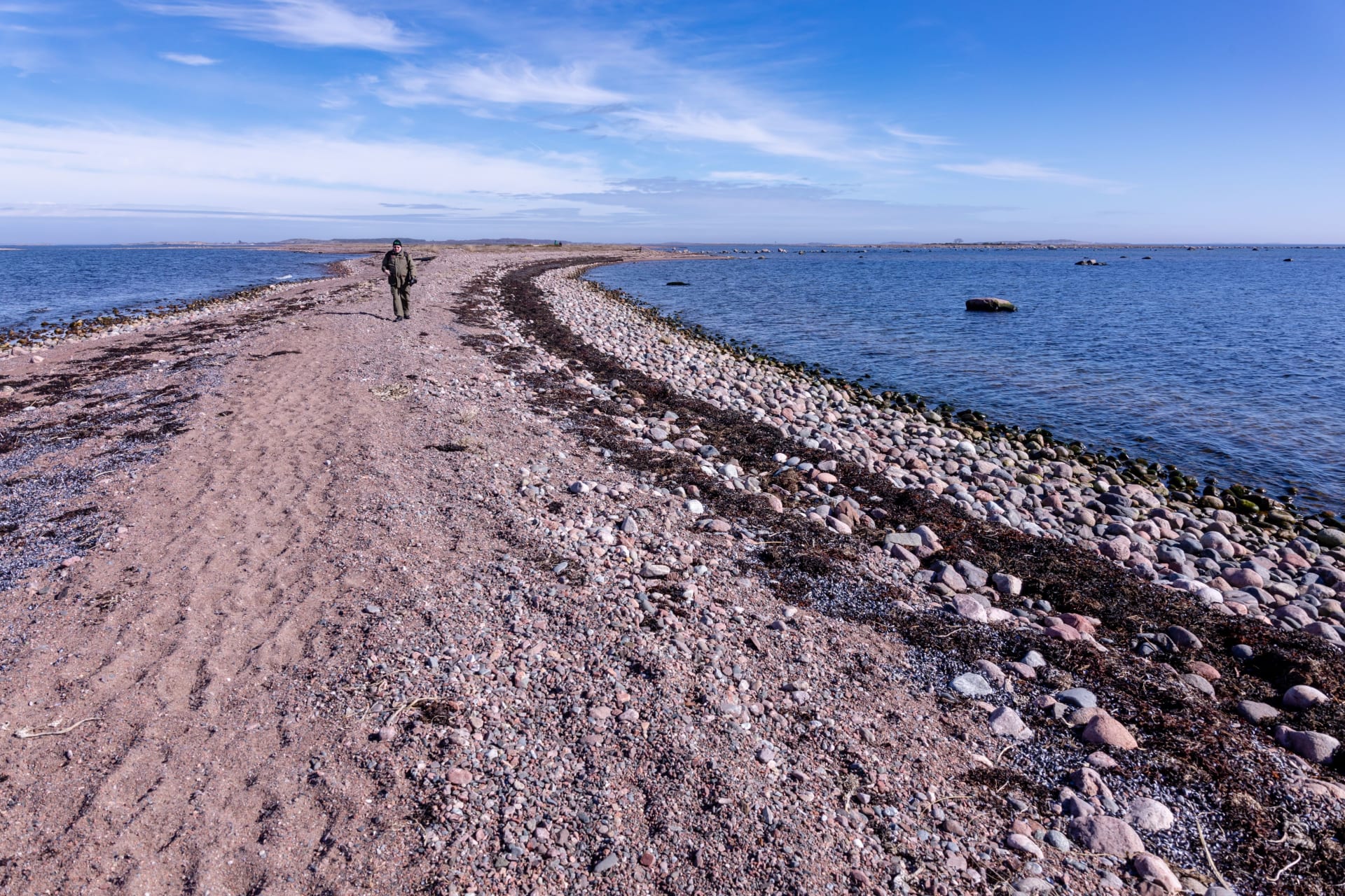 Sorasta muodostuvan kannaksen molemmin puolin on merta. Kannaksella kävelee mies.On both sides of the isthmus formed by gravel is the sea. A man is walking on the isthmus. 