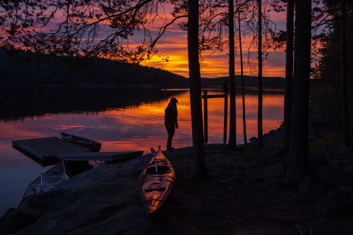 Kesäinen auringonlasku järvellä. Rannassa retkeilijä ja kajakki, taustalla järvimaisema. Kuva: Saara Lavi