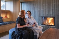 Kaksi naista nauttimassa lasit punaviiniä takkatulen äärellä Serlachius Taidesaunan olohuoneessa.