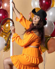 Hymyilevä nainen pirteän oranssissa asussaan pitelee ruoskaa kädessään musta stetsonhattu päässään. Taustalla ilmapalloja.