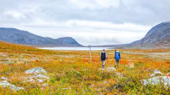 Kaksi retkeilijää syksyisessä avotunturimaisemassa Käsivarren erämaa-alueella. Taustalla suurtuntureita ja järvi. Kuva: Rami Valonen