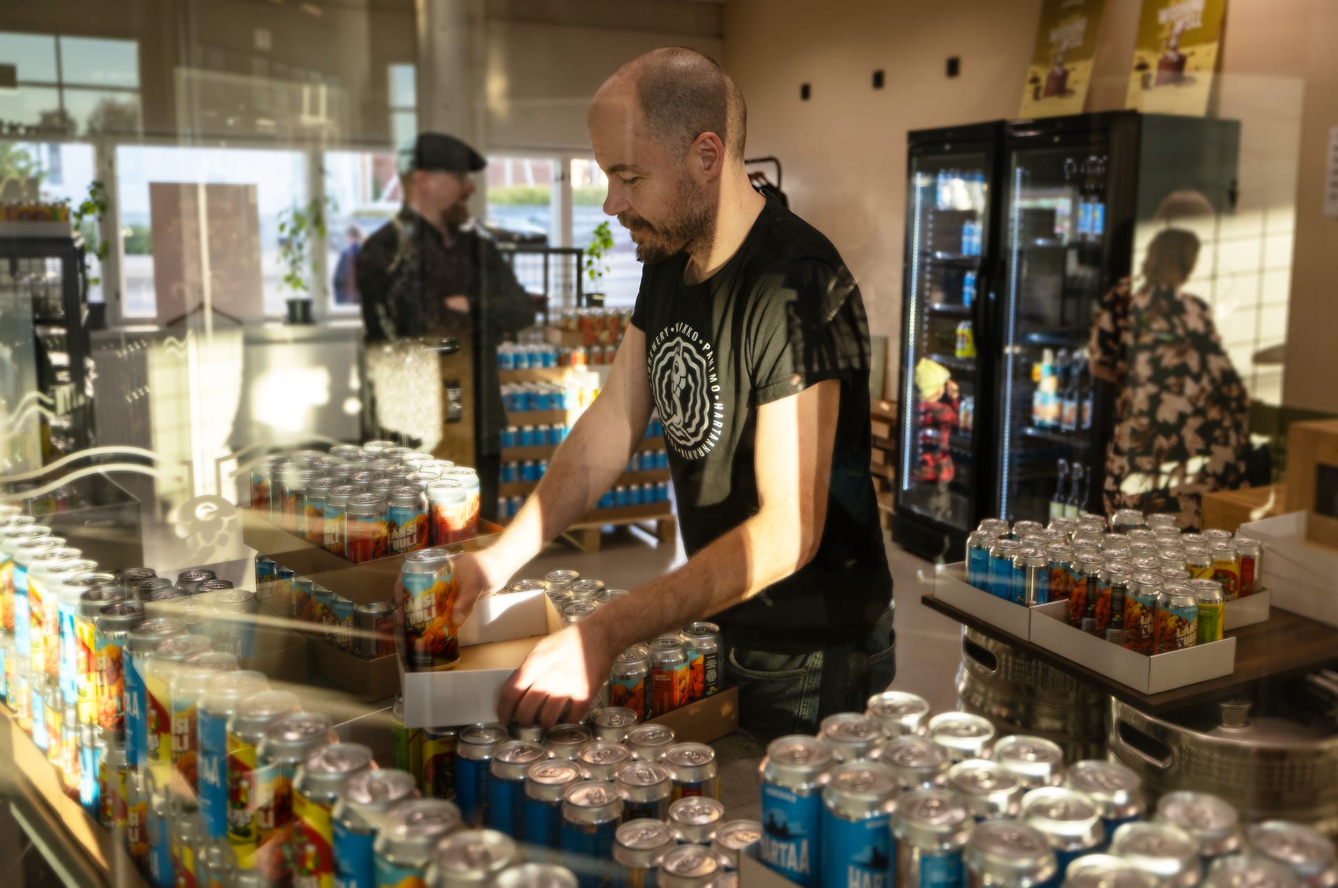 Smiling Varikko team member holding a bottle, with a wide range of beer cans on display