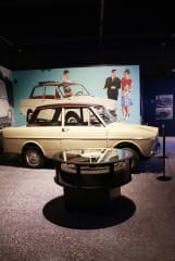 Kermanvärinen Daffodil-merkkinen auto kuvattuna sivulta. Taustalla 1960-luvun sinivoittoinen automainoskuva.