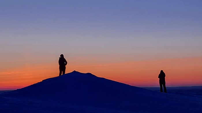 Kaksi retkeilijää katselee lumisella tunturin huipulla auringonlaskua. Tunturin taakse laskeutunut aurinko on värjännyt taivaan punertavaksi.   Two hiker is watching sunset in top of the snowy fell. Sunset makes the sky red. Photo: Petri Kulha