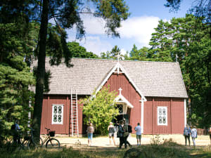 Puinen iso kirkko, jonka piha-alueella on pari polkupyörää ja ihmisiä. Kesäpäivä on kuuma.A large wooden church with a couple of bicycles and people in the yard. The summer day is hot.
