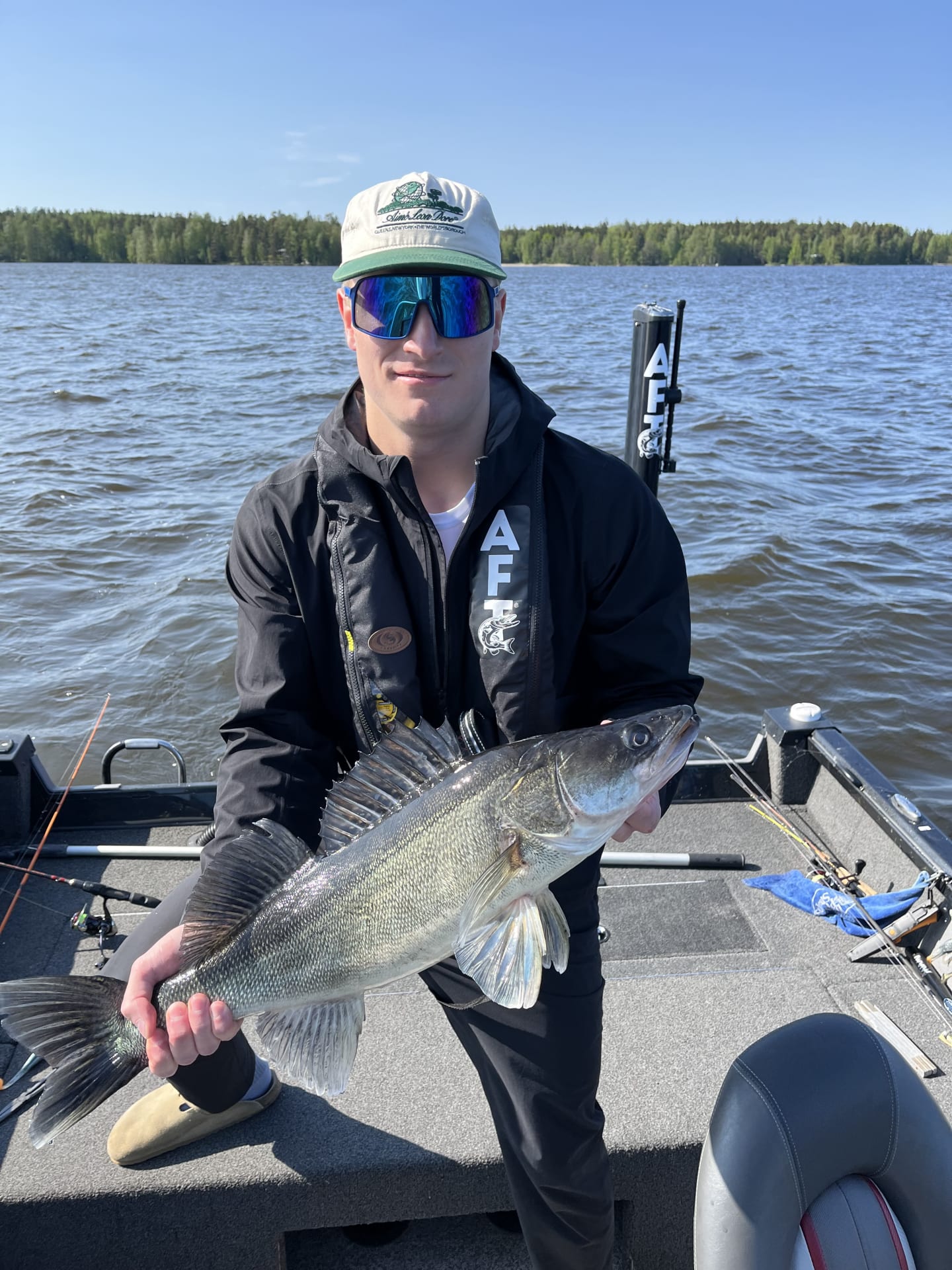 A young man holding a zander on a fishing trip. He is standing on a boat with the lake Längelmävesi behind him.