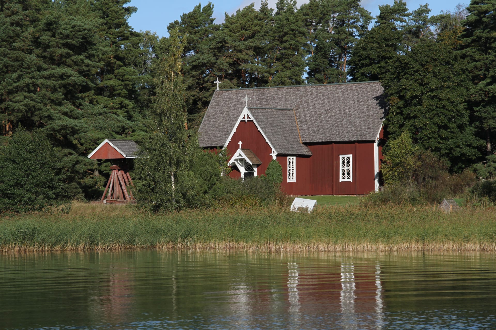 Kuva on otettu kesällä veneestä. Rannassa on ruovikkoa.  Kuivalla maalla on puinen iso kirkko. The picture was taken from a boat in the summer. There is reeds on the beach. There is a big wooden church on dry land.